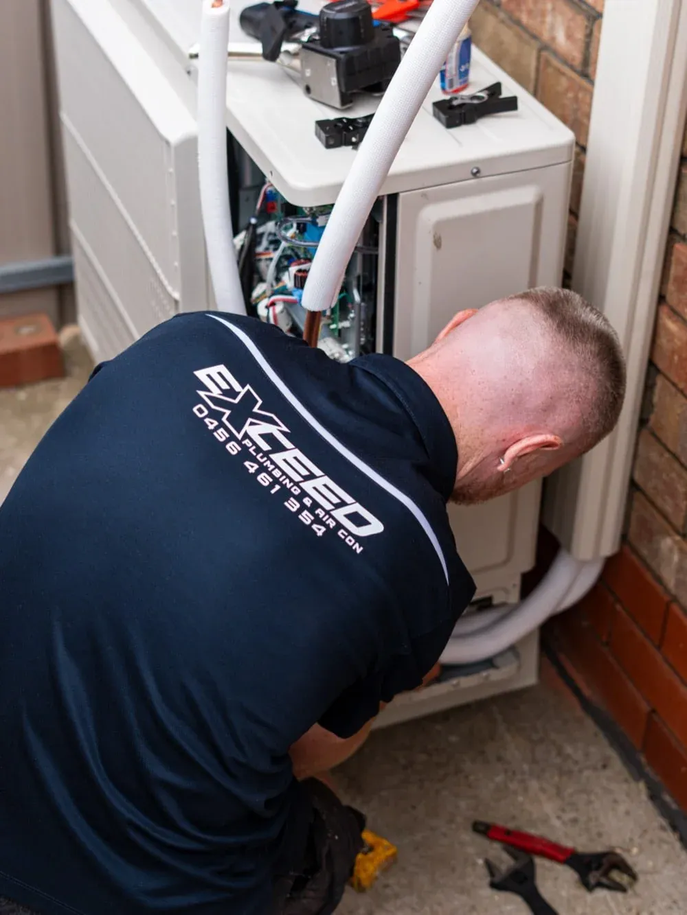Exceed Plumbing technician servicing a split system outdoor unit beside a brick wall