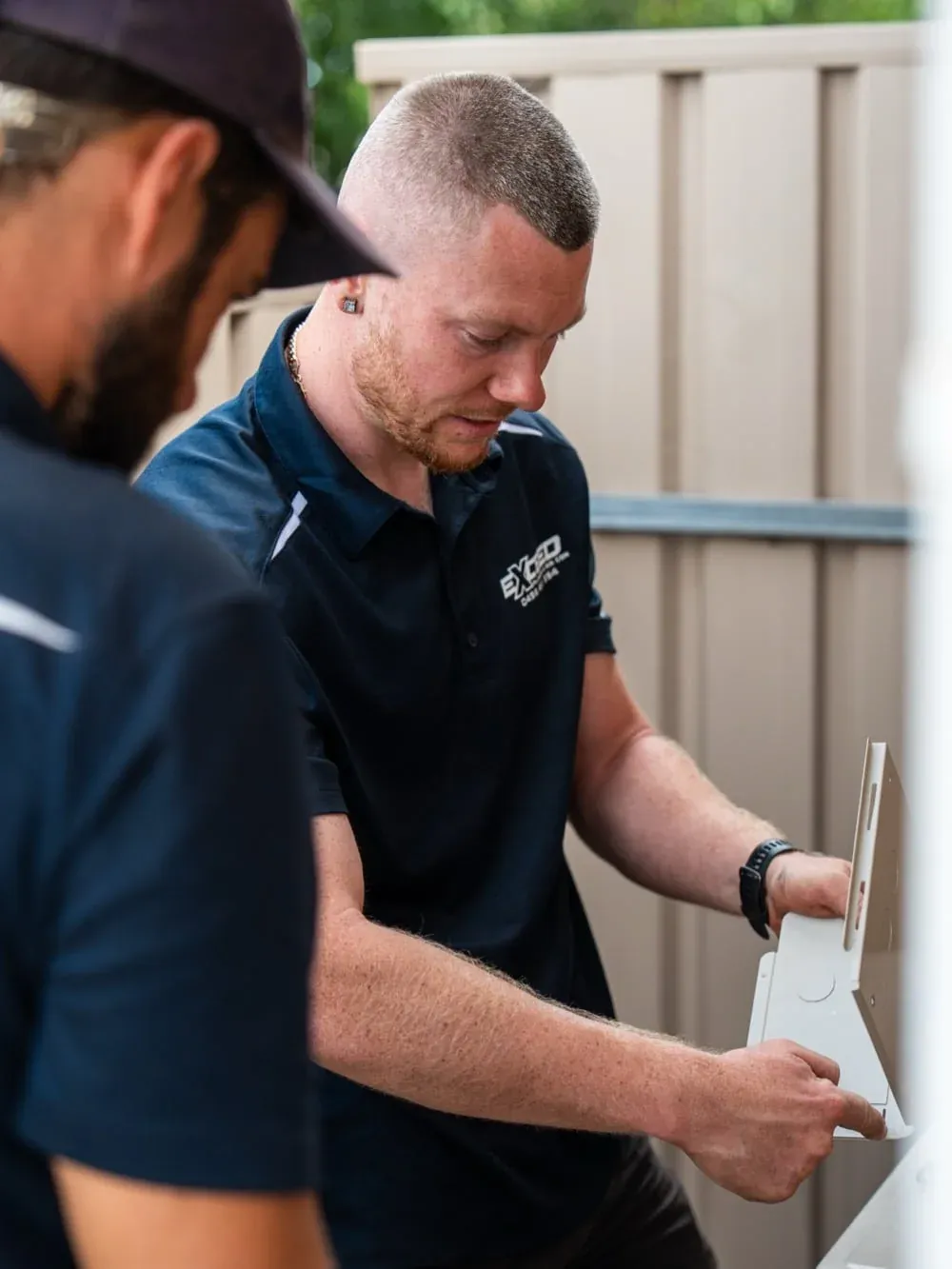 Two Exceed Plumbing technicians fitting a wall bracket for an air conditioner