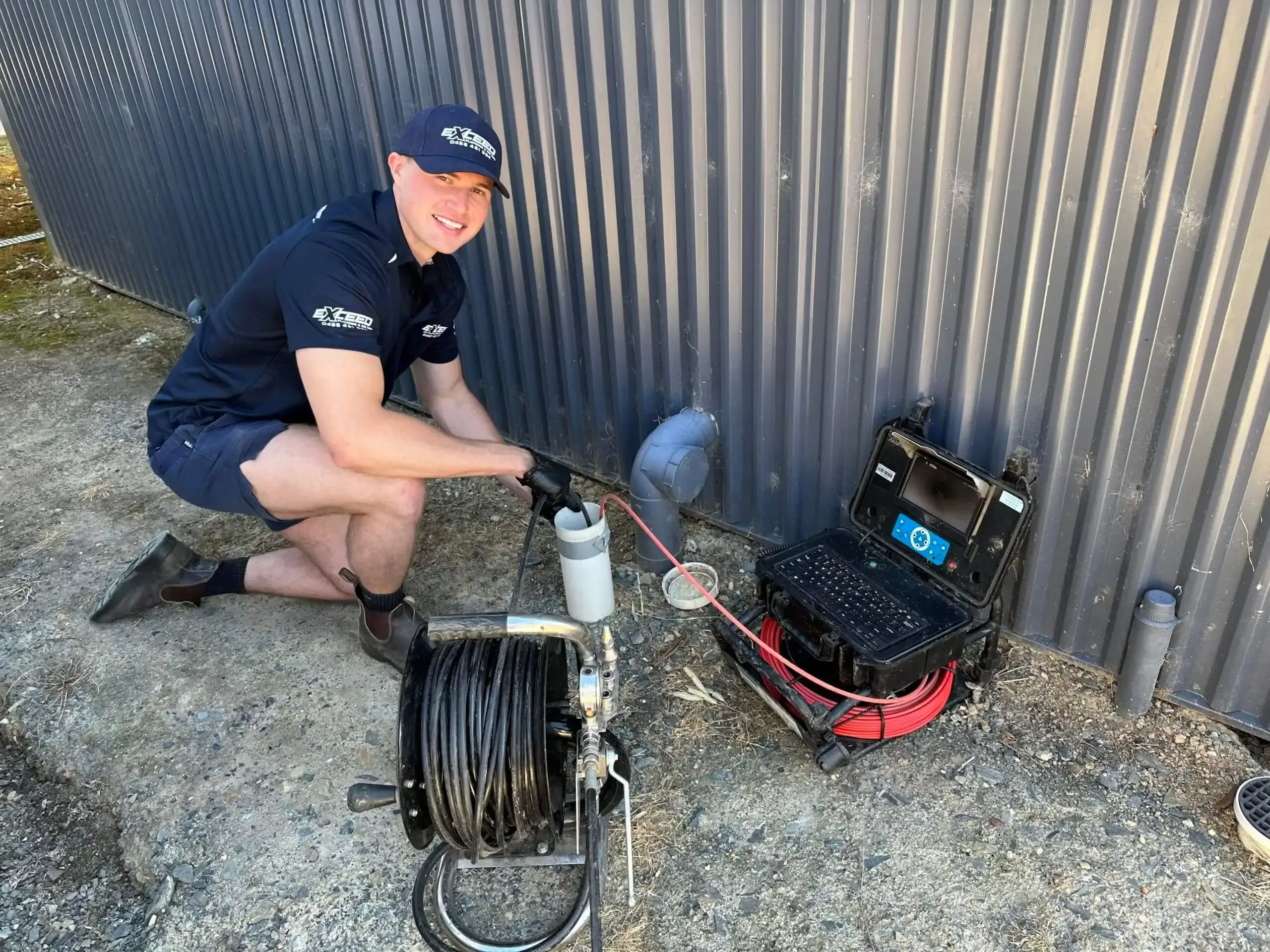 Exceed Plumbing team member jetting a blocked drain through an inspection opening, the drain camera beside him