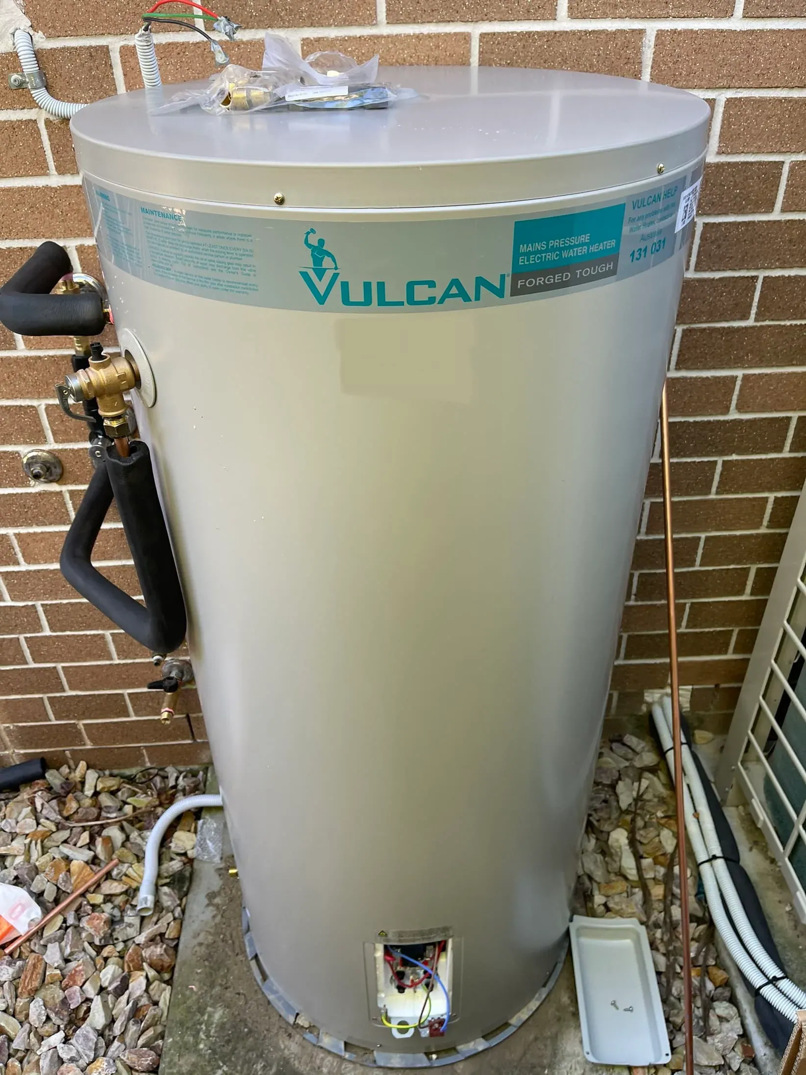 Storage hot water tank standing on gravel beside a brick wall