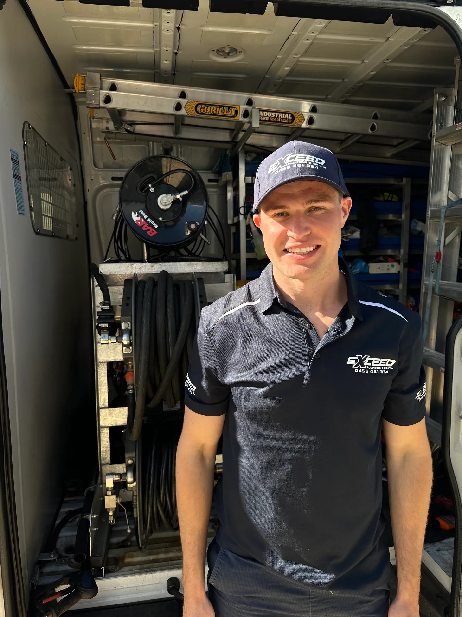 Smiling Exceed Plumbing team member at the back of the service van, the drain jetter’s hose reel behind him