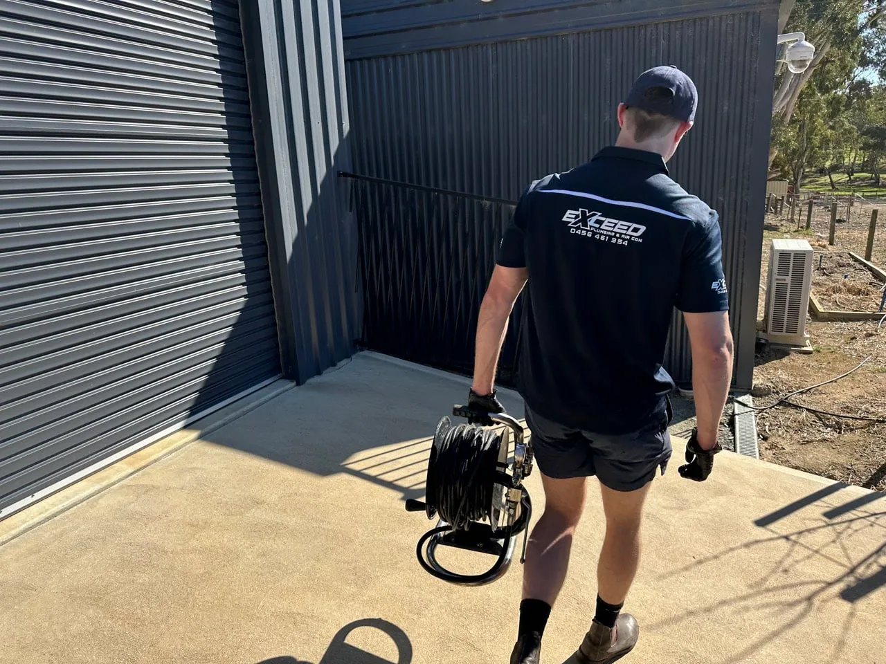 Exceed Plumbing plumber carrying a drain camera reel beside a shed