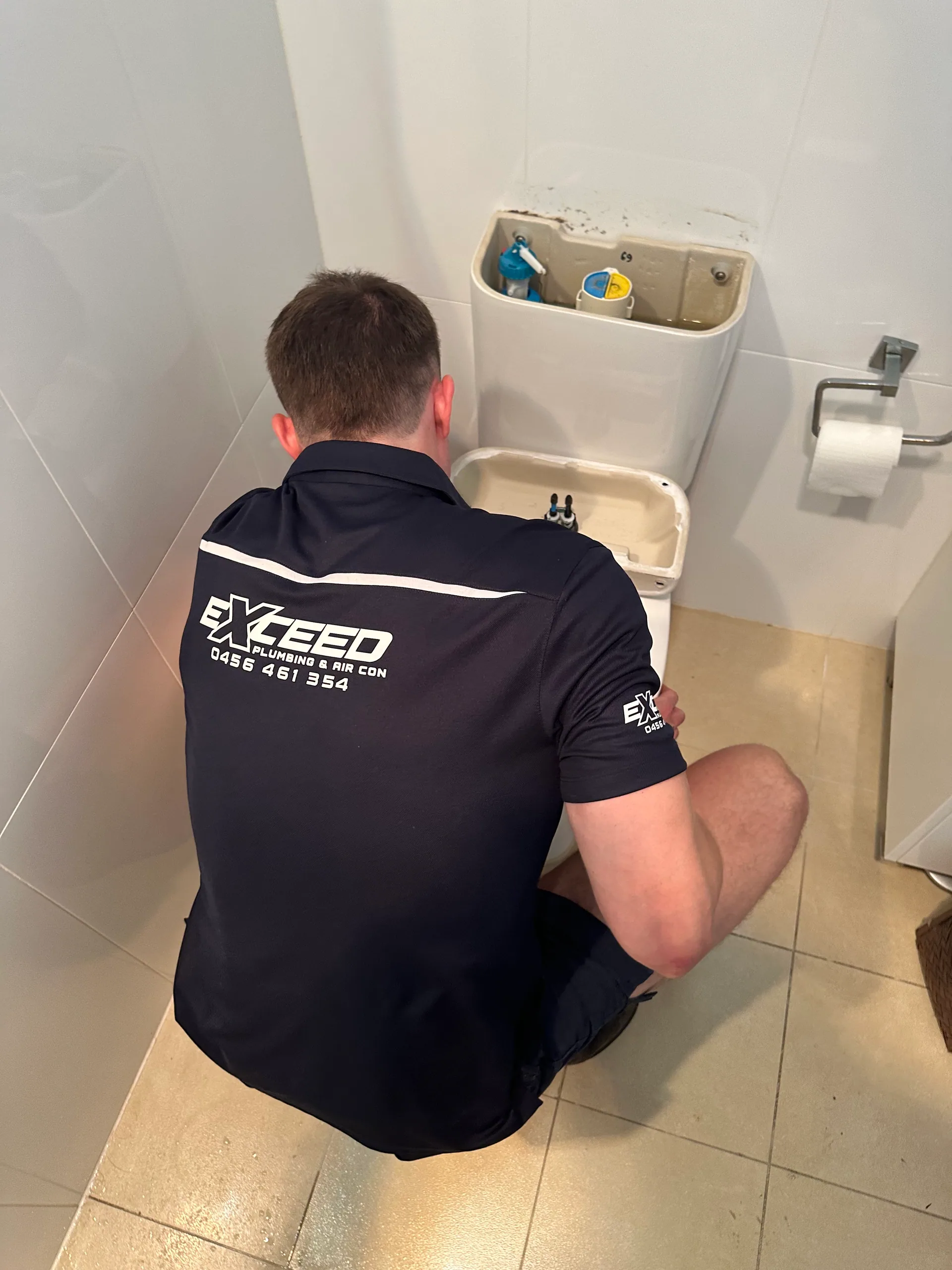 Exceed Plumbing plumber kneeling at a toilet with the cistern lid off