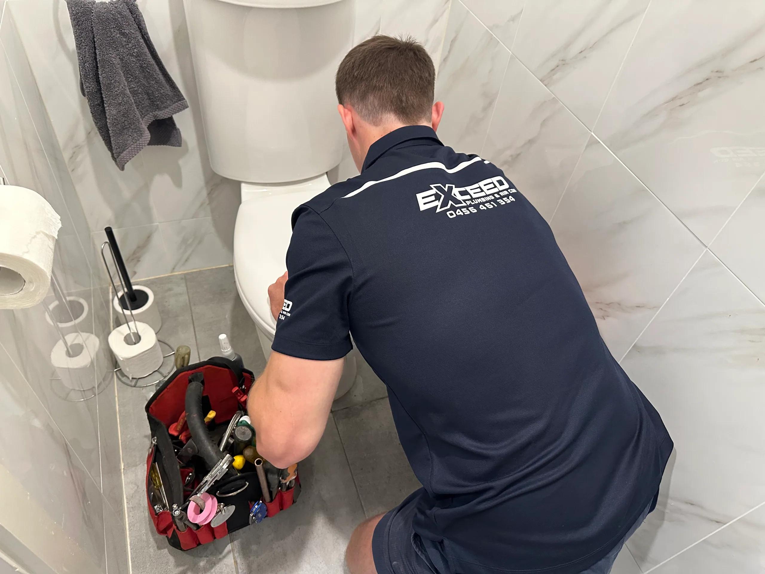 Exceed Plumbing plumber working on a toilet beside his toolbag