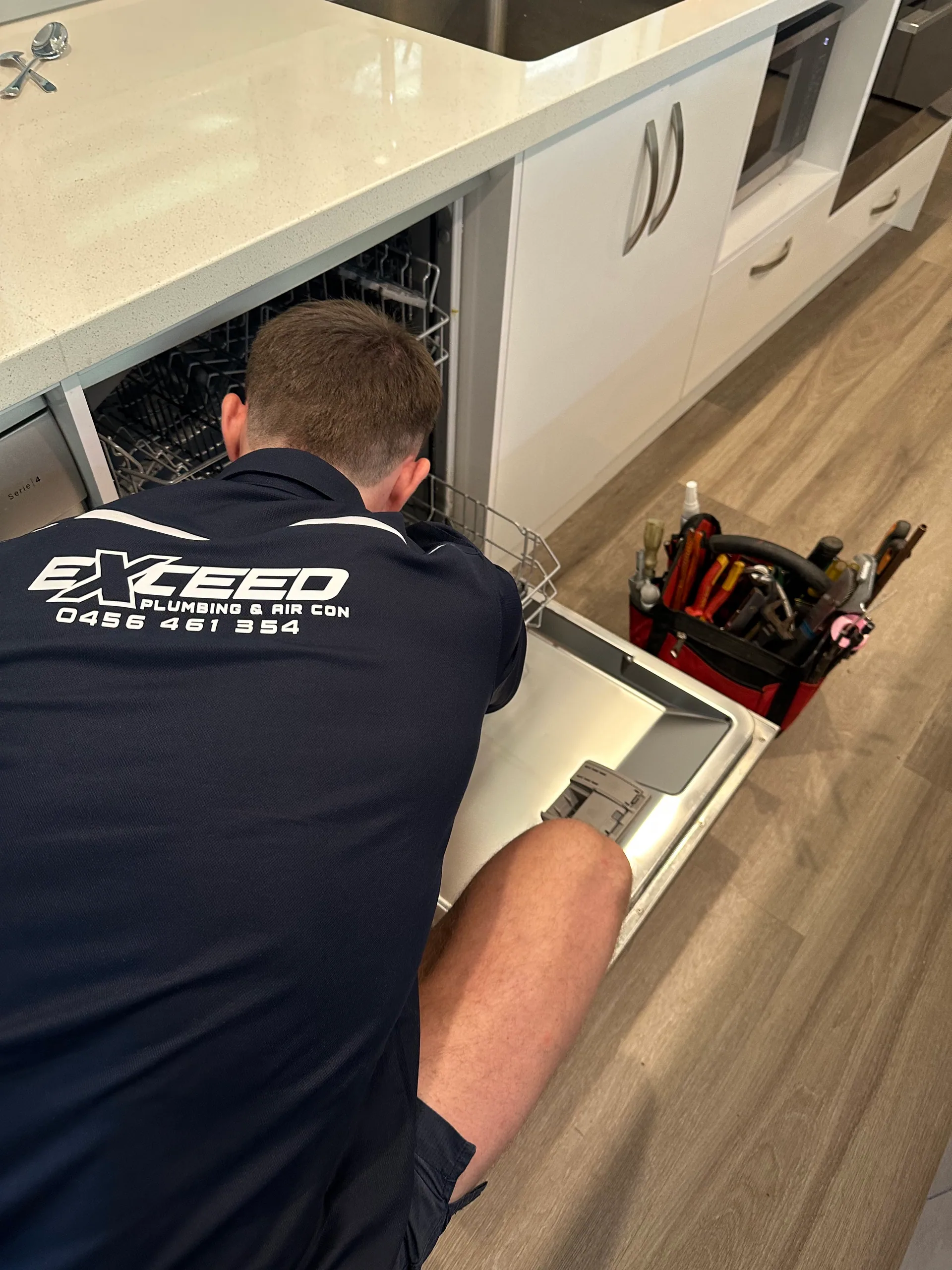 Exceed Plumbing plumber reaching into a kitchen cabinet beside a dishwasher