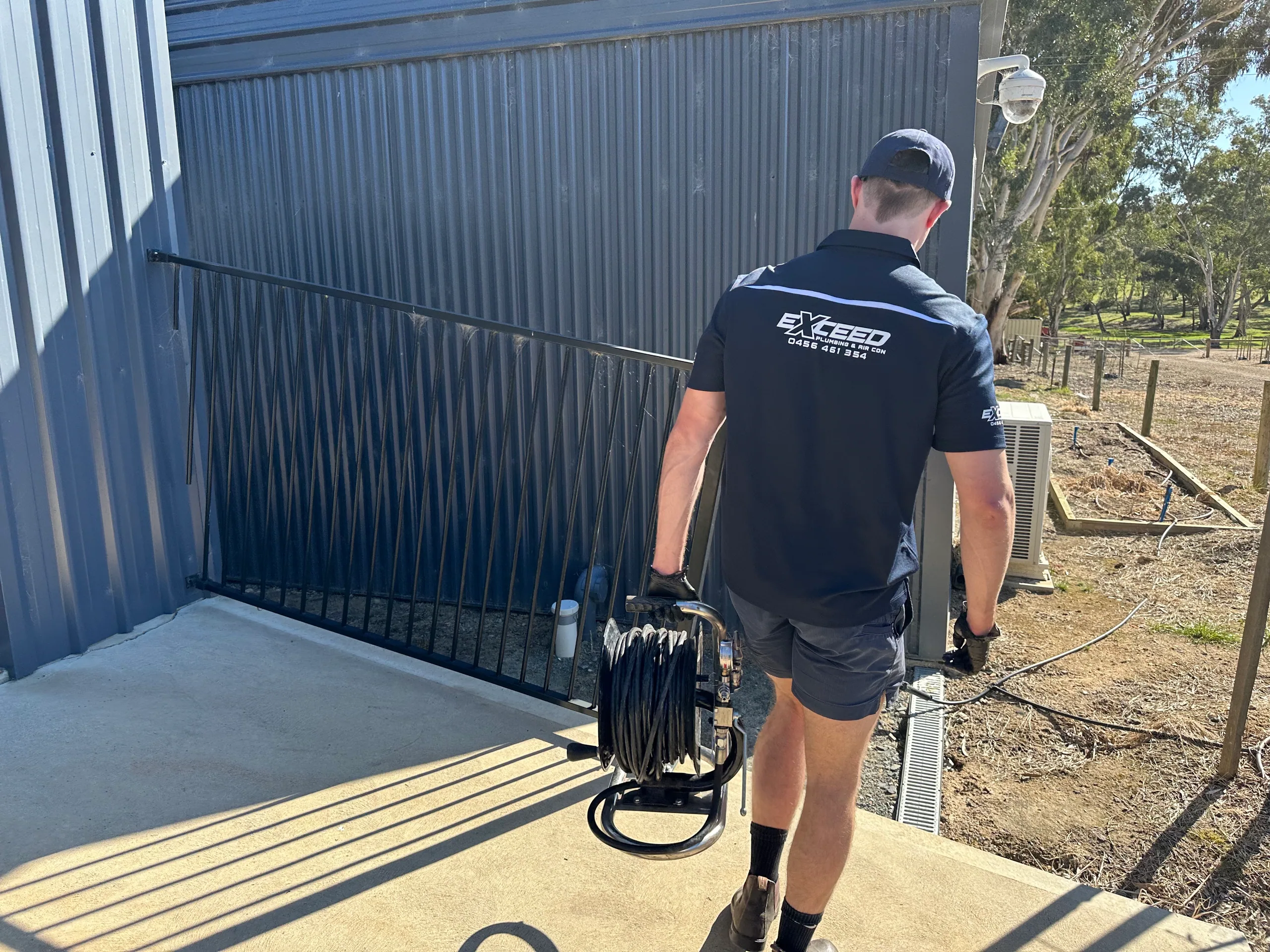 Exceed Plumbing plumber carrying a drain camera reel along a shed