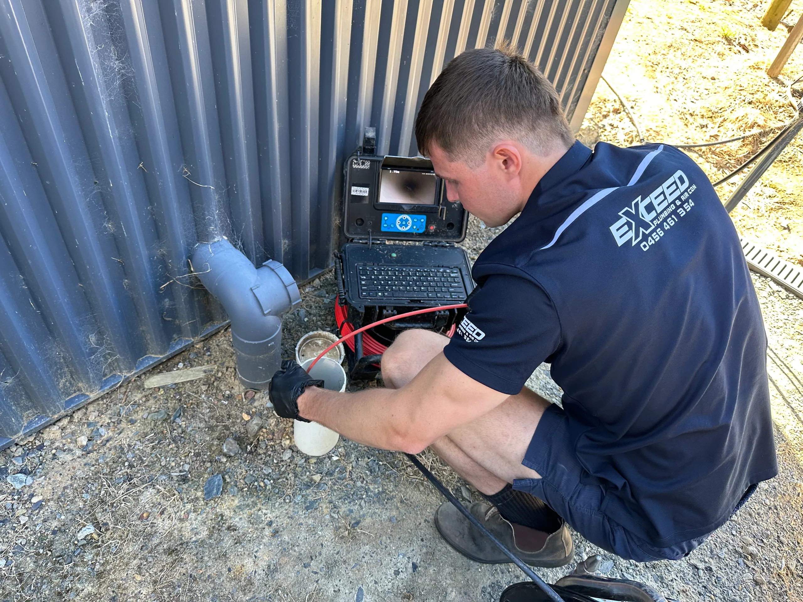 Exceed Plumbing plumber feeding a drain camera into a pipe beside a shed