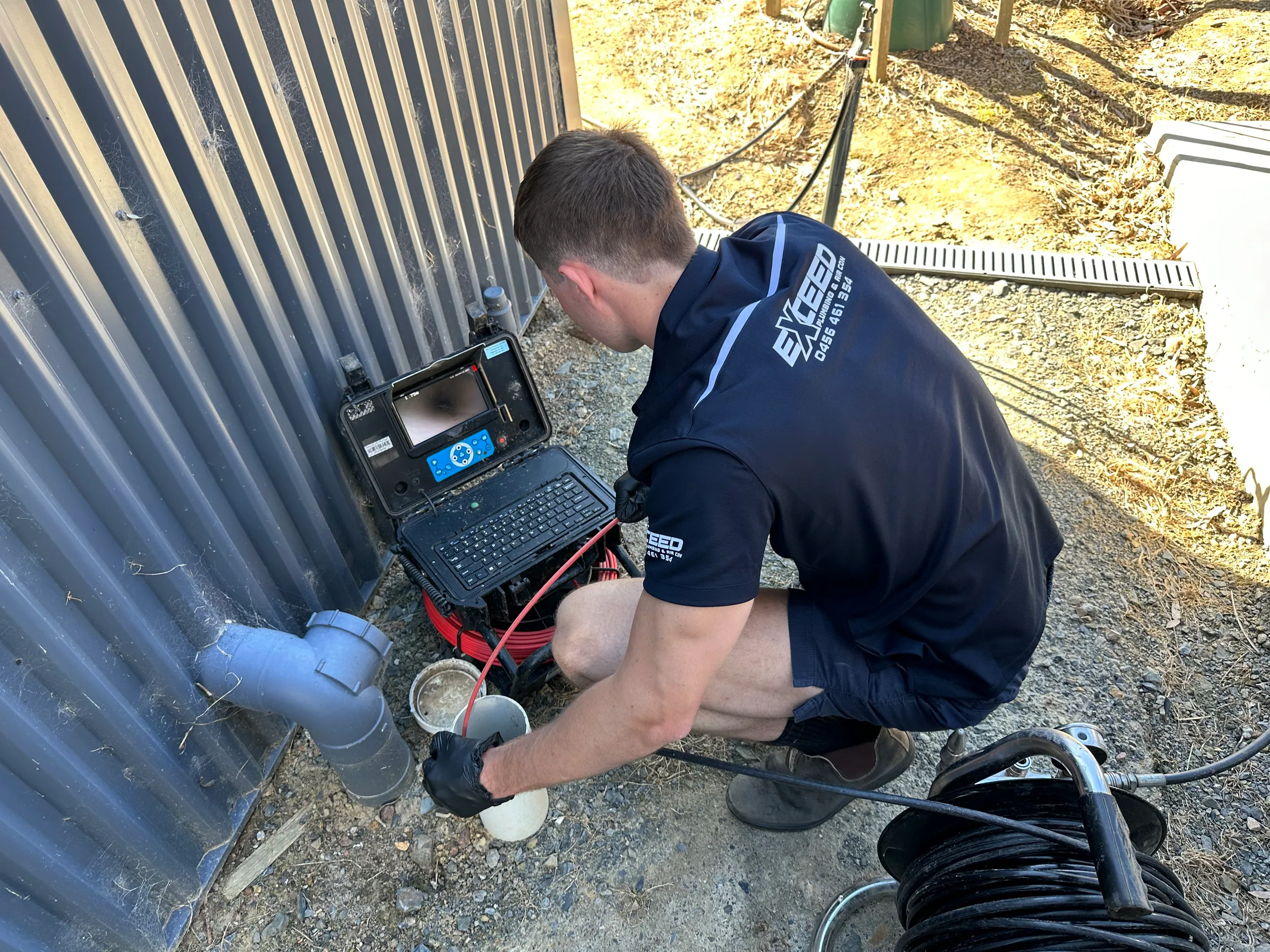 Exceed Plumbing plumber crouched at a drain camera beside a shed