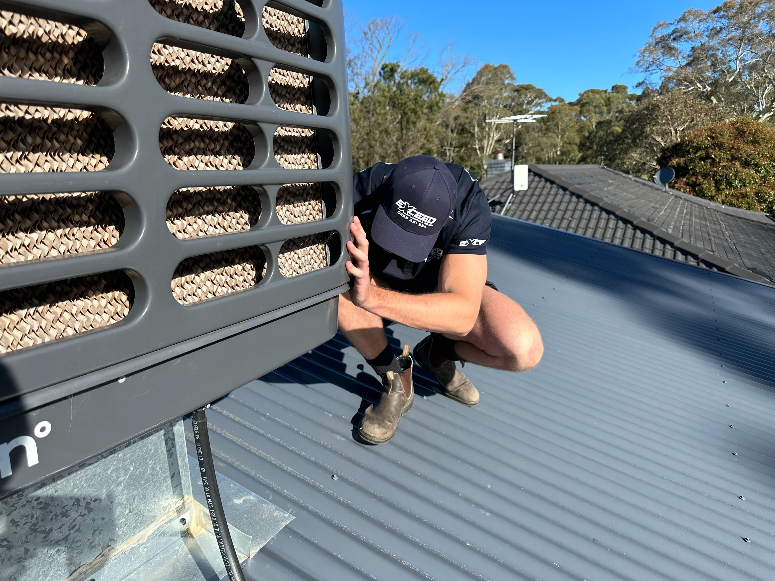 Exceed Plumbing plumber crouching beside an evaporative cooler on a metal roof