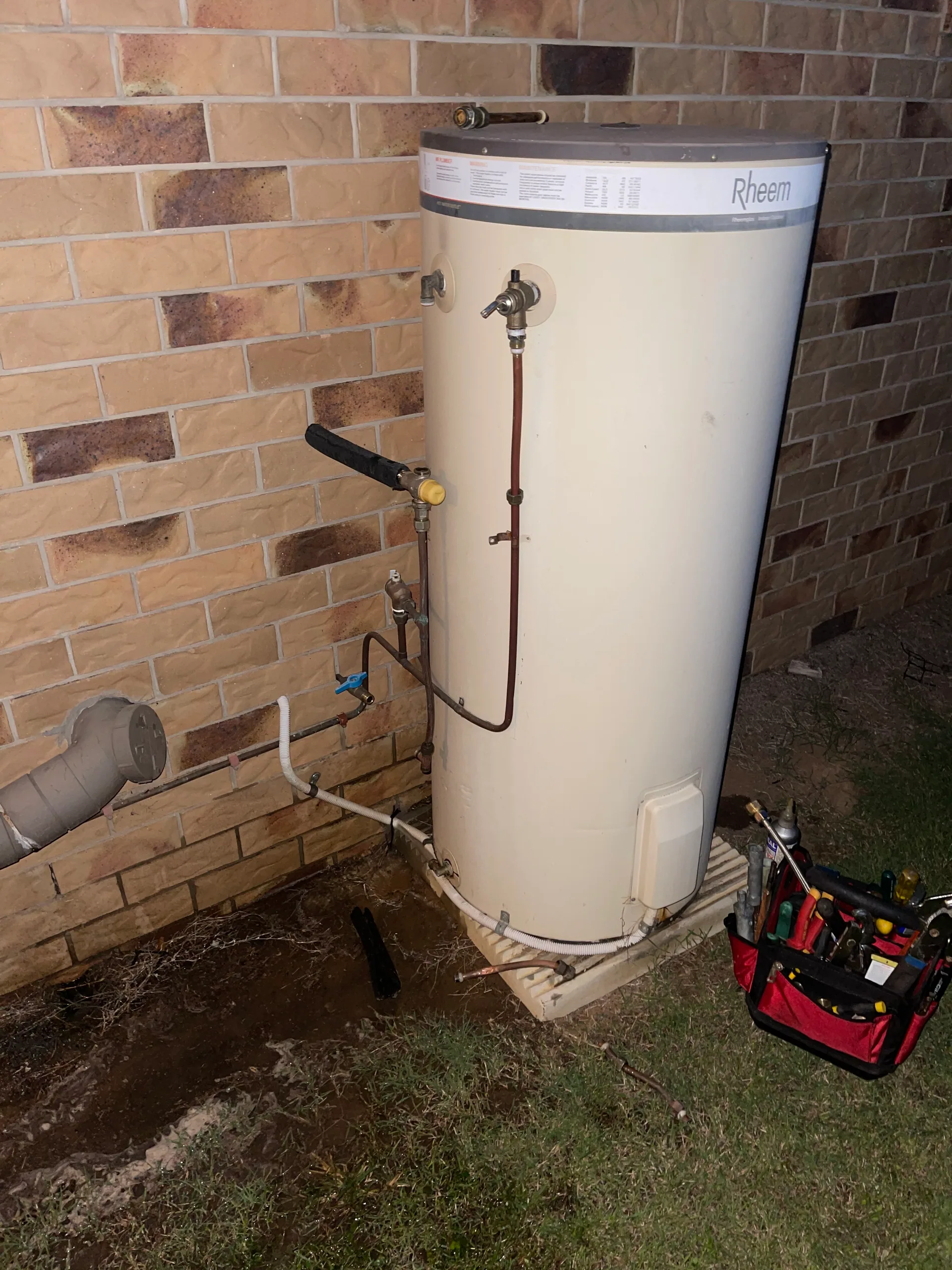 Rheem hot water system against a brick wall beside a plumber's toolbag