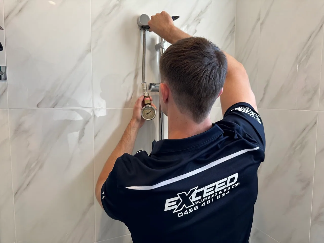 Exceed Plumbing plumber fitting a shower rail on a marble-look tiled wall