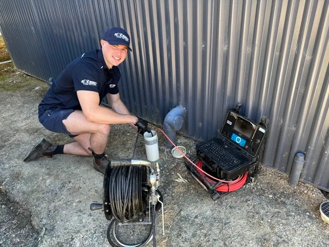 Exceed Plumbing plumber setting up a drain camera beside a shed