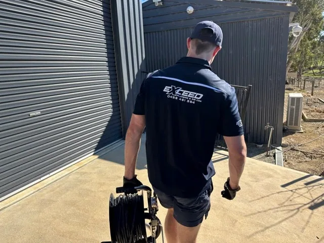 Exceed Plumbing plumber carrying a drain camera reel past a roller door