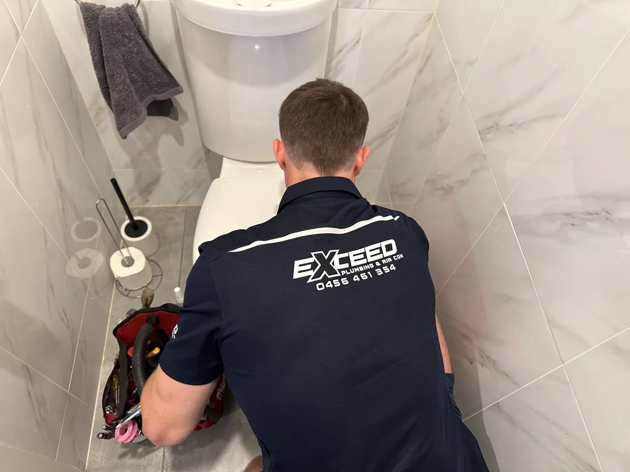 Exceed Plumbing plumber kneeling at a toilet with his toolbag